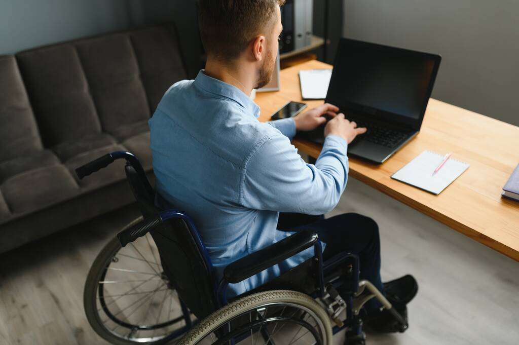 Młody szczęśliwy przedsiębiorca na wózku inwalidzkim pracujący na laptopie w domu. - Zdjęcie, obraz