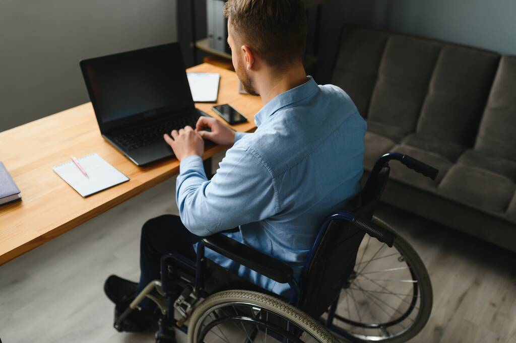 Людина з обмеженими можливостями в інвалідному візку працює в офісі за комп'ютером. Він посміхається і захоплюється робочим процесом
. - Фото, зображення