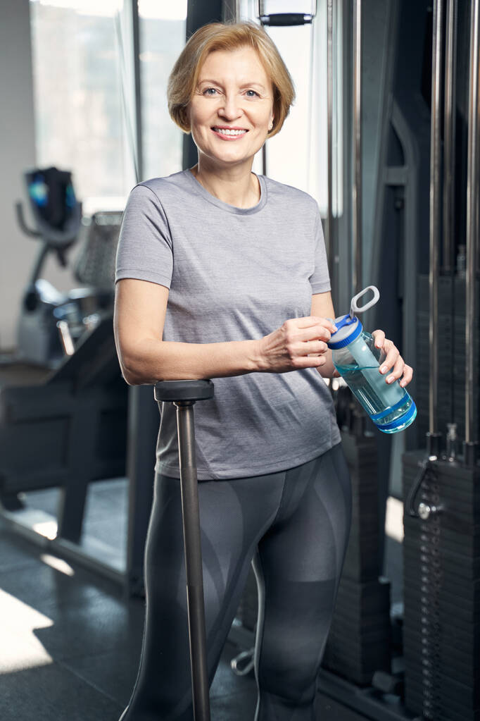 Mujer alegre con botella de agua de pie en el gimnasio - Foto, imagen