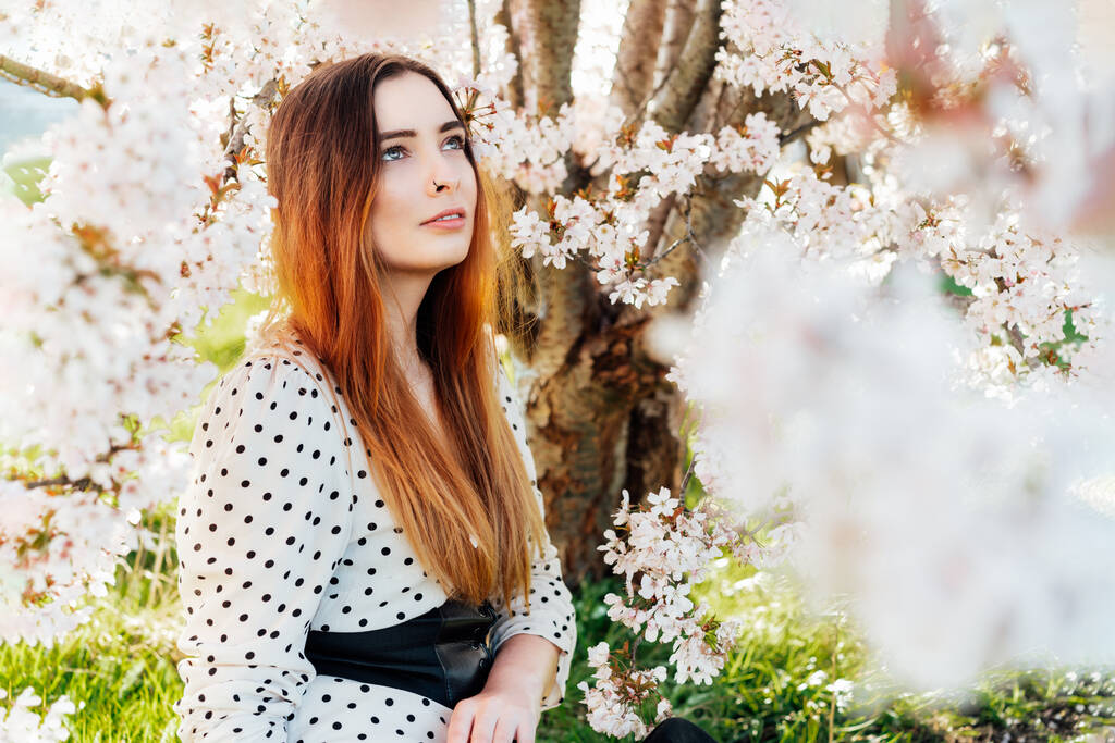 ピンクの花の木の近くに座って太陽を楽しむドレスで若いブルネットの女性の肖像画。Z世代の女の子は晴れた日の間に春の気分をお楽しみください。自然の中でリラックス. - 写真・画像