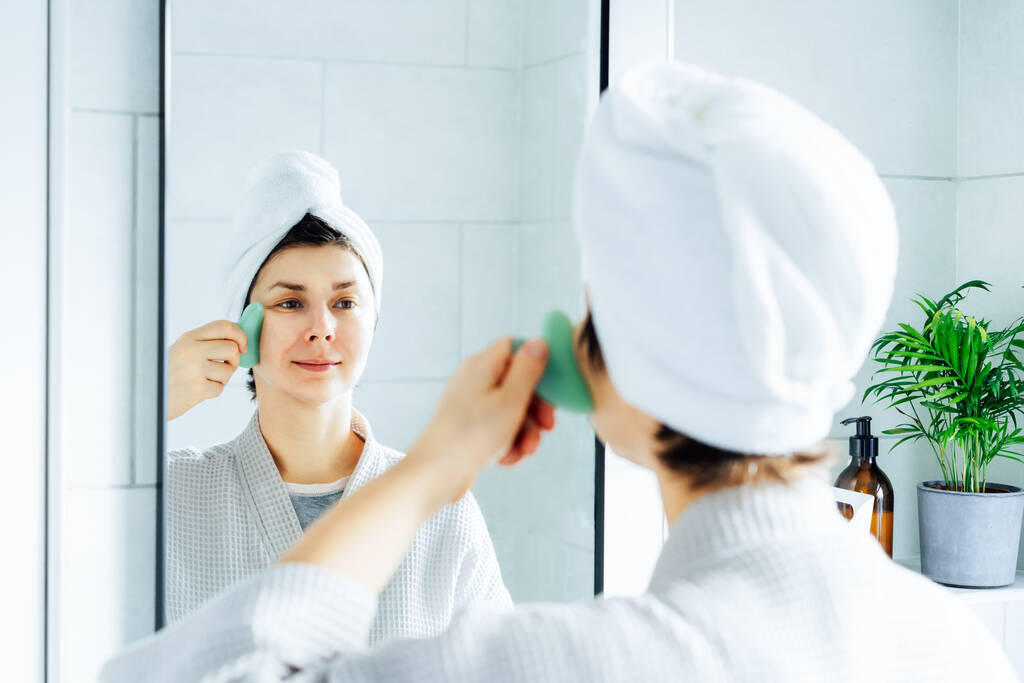 Женщина в халате с полотенцем на голове делает массаж лица гуа ша роллером в ванной комнате. Понятие естественных процедур по уходу за кожей. Домашняя косметология. Селективный фокус. - Фото, изображение