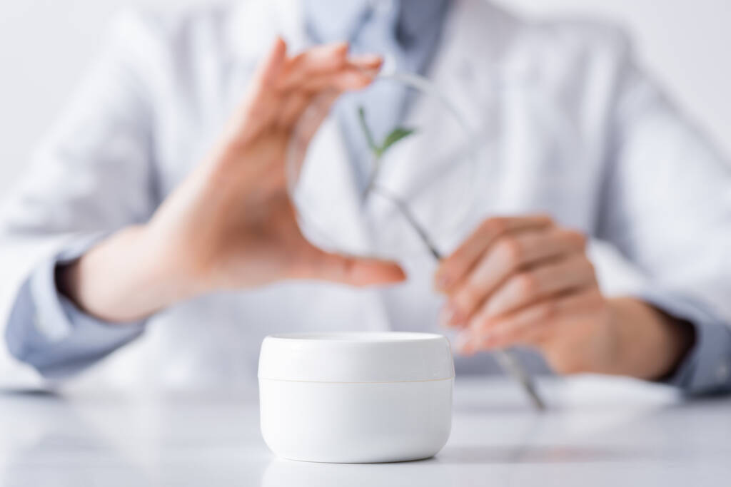 container with cream near blurred laboratory assistant in lab - Photo, Image