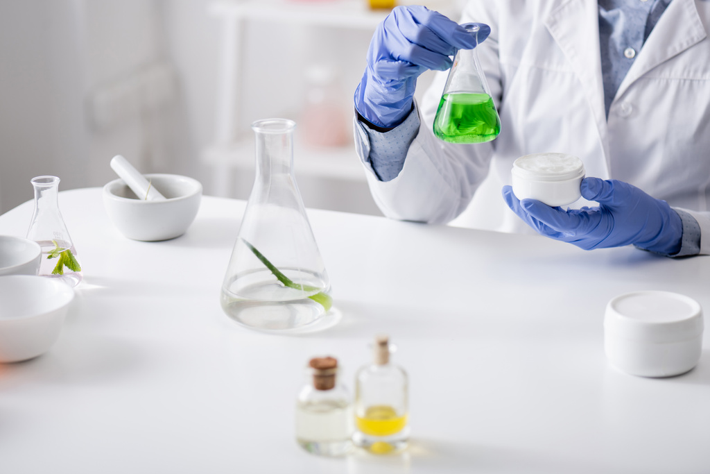 visão recortada de assistente de laboratório em luvas de látex segurando frasco com líquido verde e creme em recipiente  - Foto, Imagem