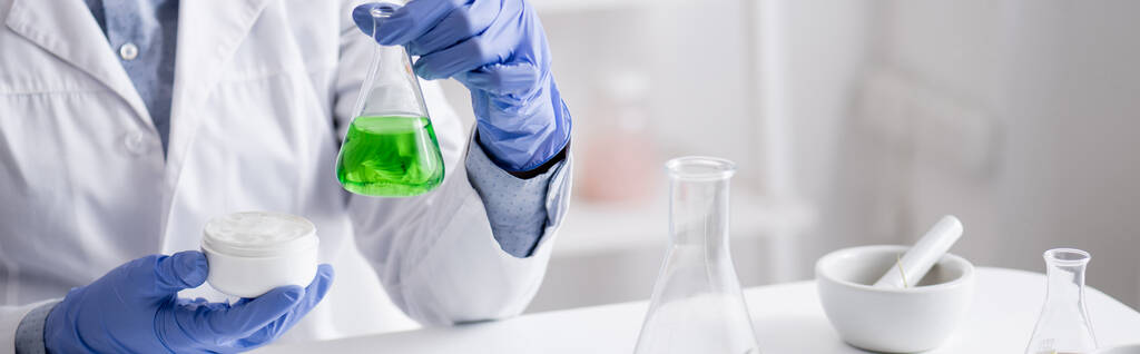 Lateks eldivenli laboratuvar asistanının yeşil sıvı ve kremalı matara ve pankart tutuşu. - Fotoğraf, Görsel
