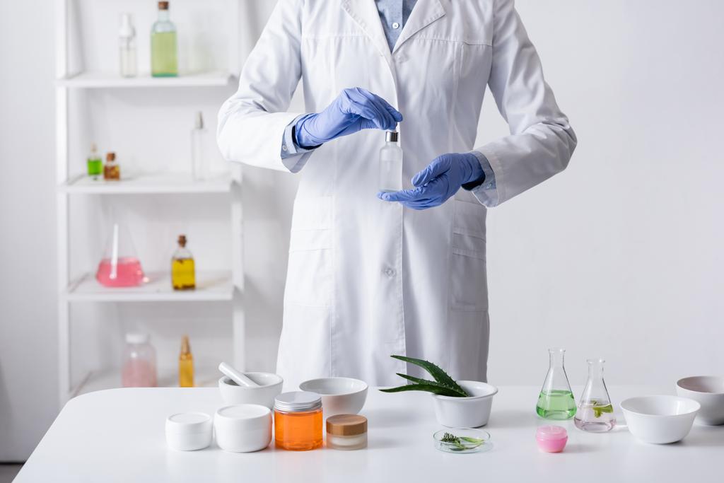 przycięty widok asystenta laboratoryjnego w rękawiczkach lateksowych trzymających butelkę z surowicą w laboratorium - Zdjęcie, obraz