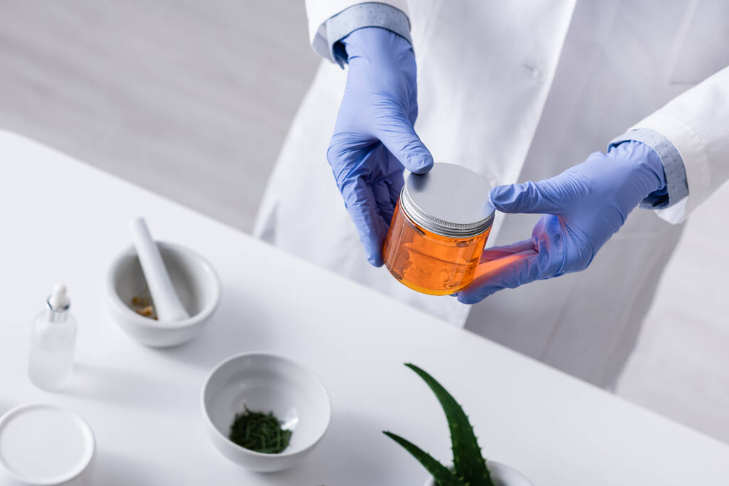 προβολή υψηλής γωνίας του εργαστηριακού βοηθού σε γάντια λατέξ κρατώντας βάζο με μέλι στο εργαστήριο - Φωτογραφία, εικόνα