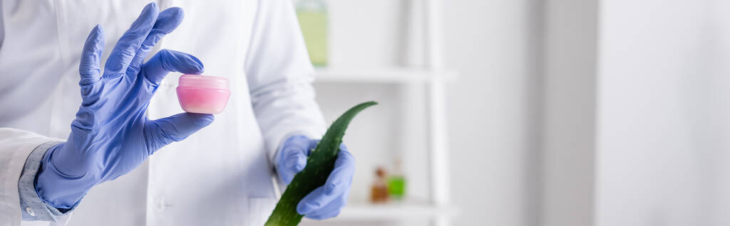 cropped view of laboratory assistant in latex gloves holding aloe leaf and container with cream, banner - Photo, Image