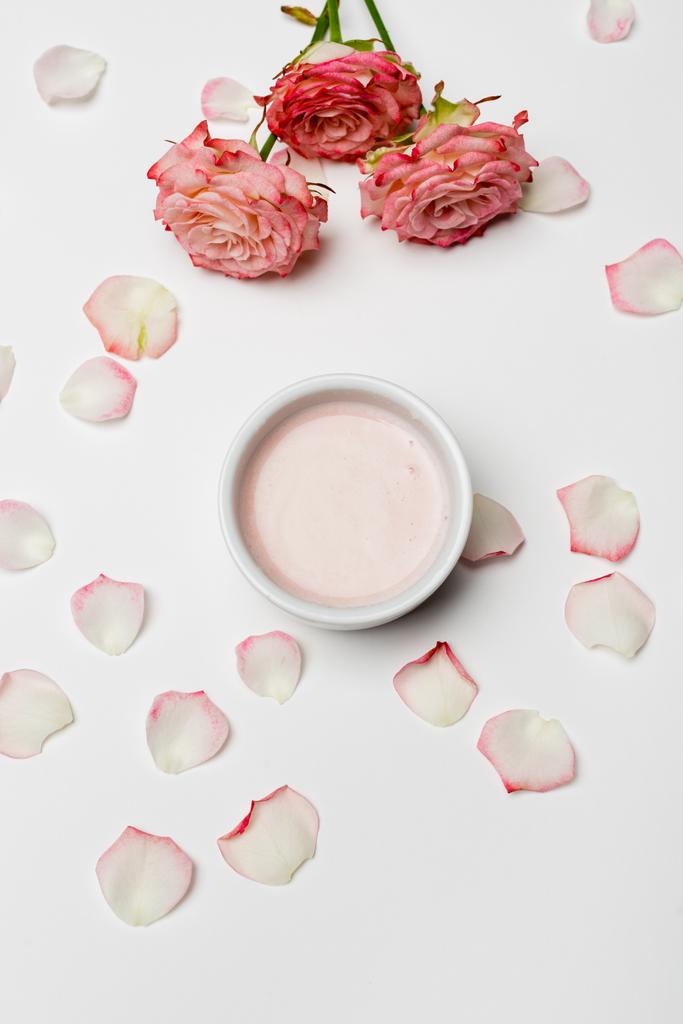 vrchní pohled na růžové květy a okvětní lístky v blízkosti mísy se smetanou na bílém - Fotografie, Obrázek