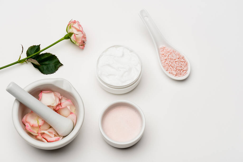 vista dall'alto del fiore vicino al sale marino rosa, contenitore con crema, pestello e malta con petali su bianco - Foto, immagini