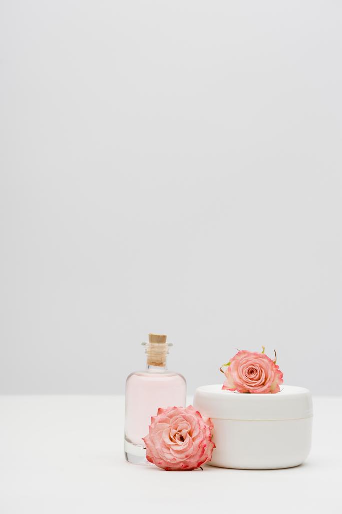 botella con extracto cerca de contenedor con crema y flores de color rosa sobre blanco - Foto, Imagen