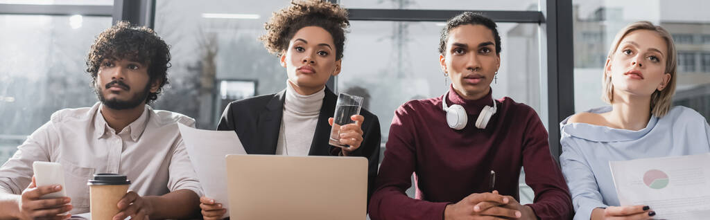 Gente de negocios interraciales mirando hacia otro lado cerca de papeles, computadora portátil y café en la oficina, pancarta  - Foto, Imagen