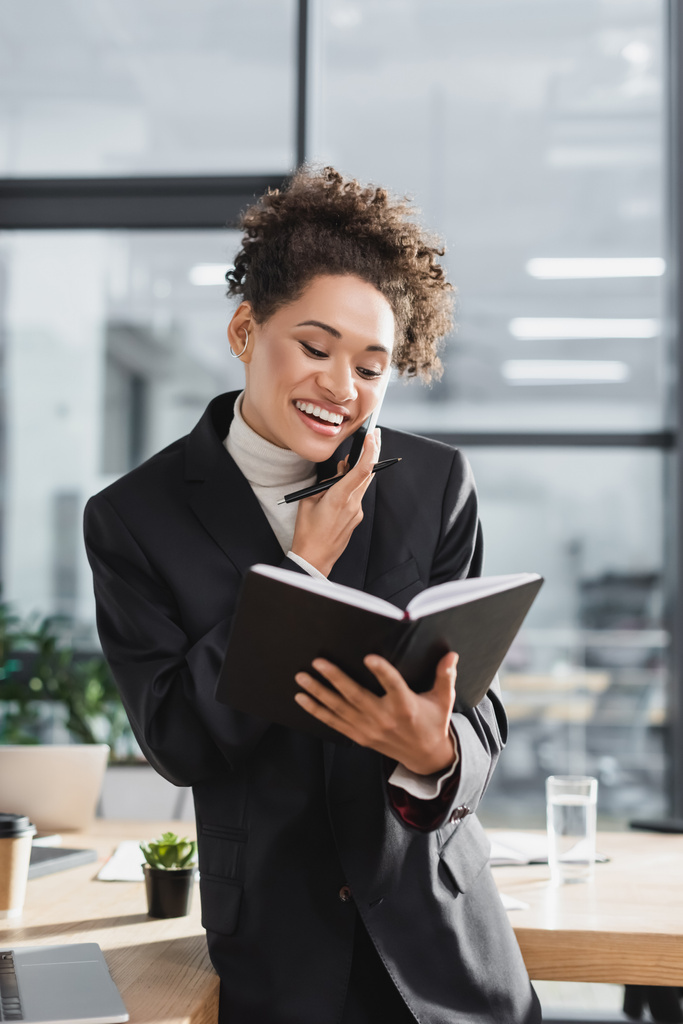 Empresaria afroamericana positiva sosteniendo un cuaderno y hablando en el teléfono inteligente en la oficina  - Foto, imagen