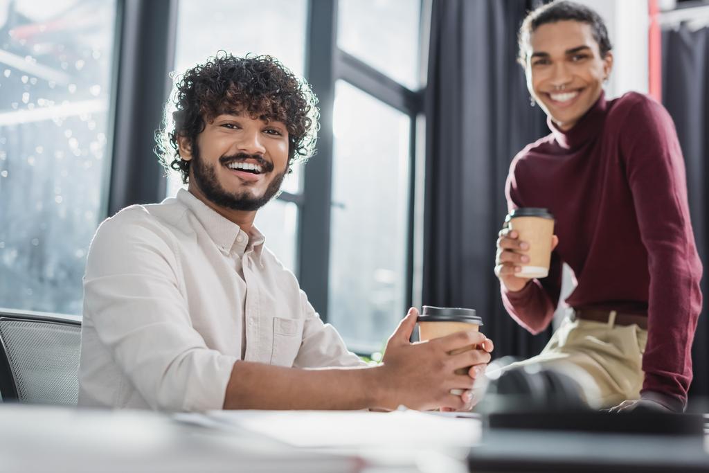 Cheerful indian businessman holding coffee to go near blurred african american colleague in office  - 写真・画像