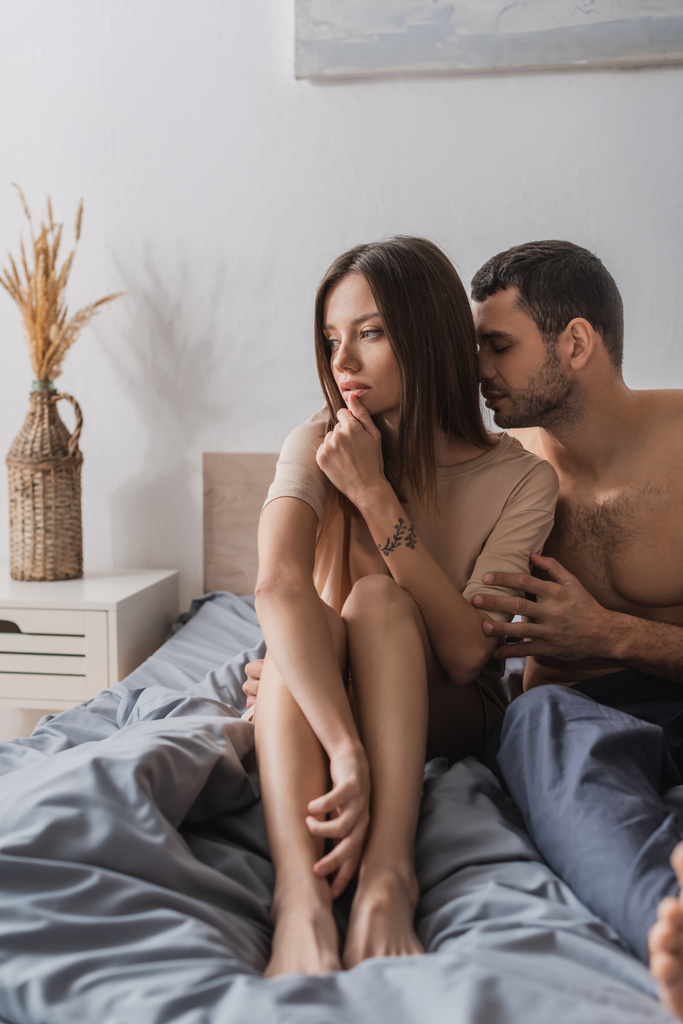 Σέξι άντρας φιλάει σκεπτική κοπέλα με μπλουζάκι στο κρεβάτι στο σπίτι  - Φωτογραφία, εικόνα