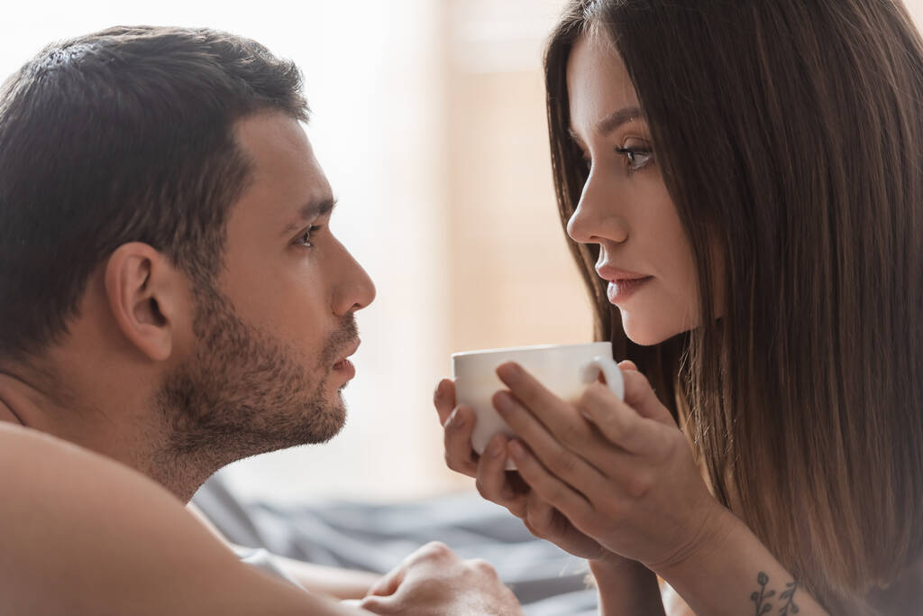 Young woman holding cup of coffee and looking at boyfriend in bedroom  - Photo, Image