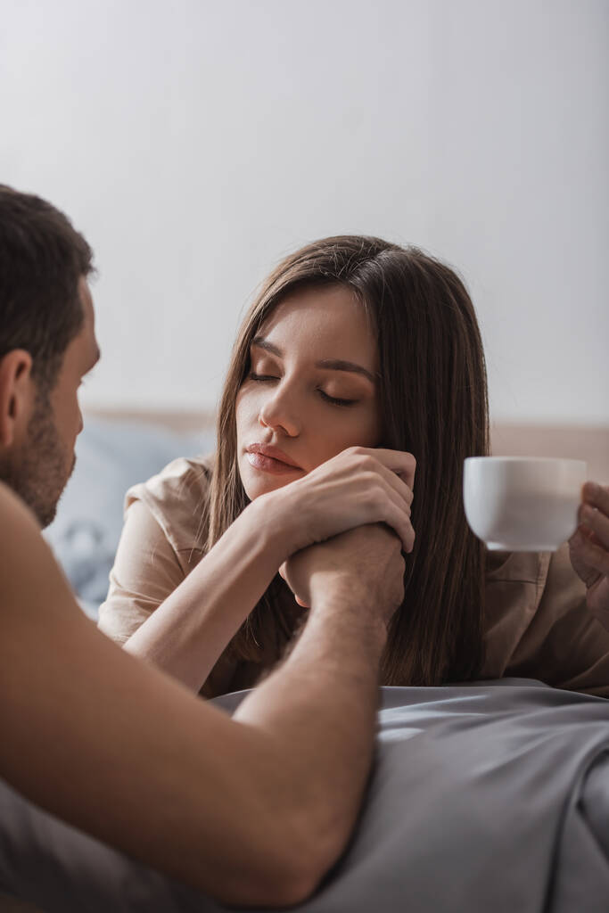 homme flou tenant la main de jolie petite amie avec tasse de café sur le lit  - Photo, image