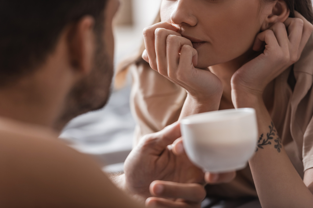 Vágott kilátás tetovált nő feküdt az ágyon közelében elmosódott férfi egy csésze kávét otthon  - Fotó, kép