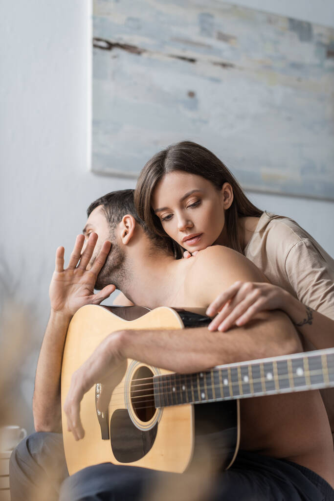 Jolie femme touchant bras de copain torse nu tenant guitare acoustique à la maison  - Photo, image