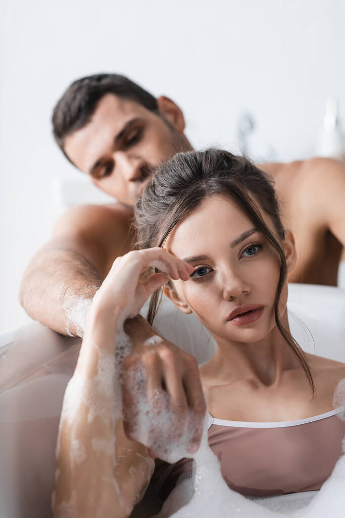 Jeune femme prenant un bain avec de la mousse près de petit ami flou à la maison  - Photo, image