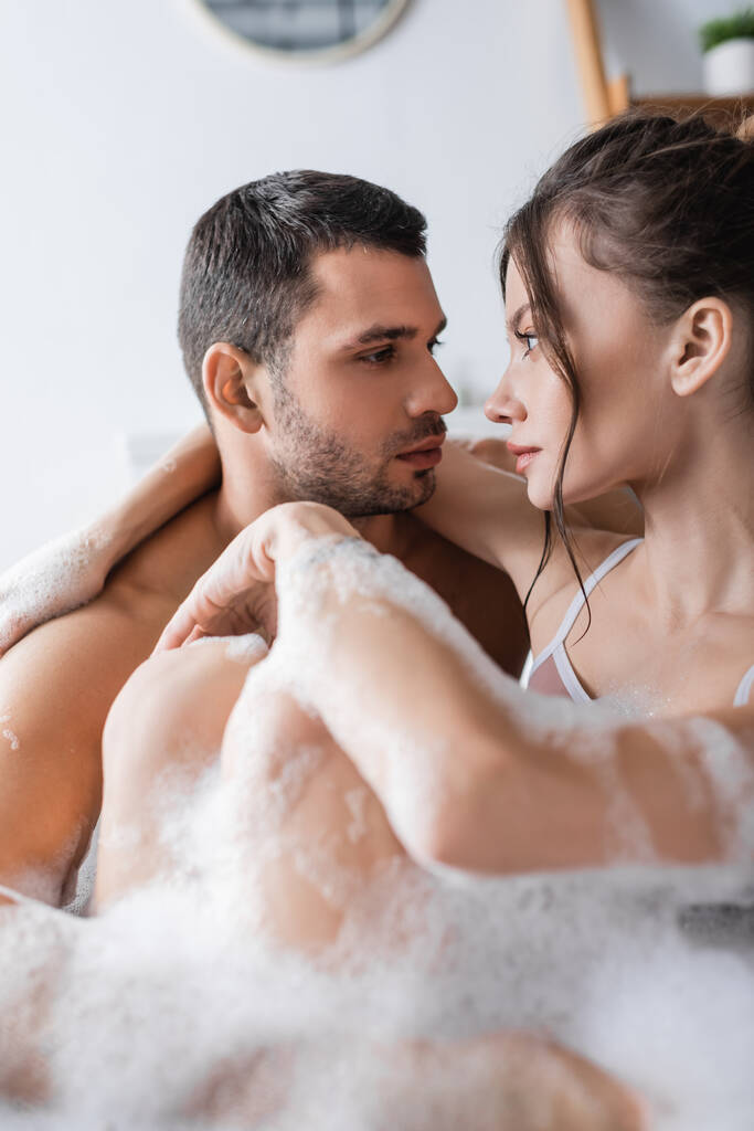 Молодая пара смотрит друг на друга, принимая ванну с пеной дома  - Фото, изображение