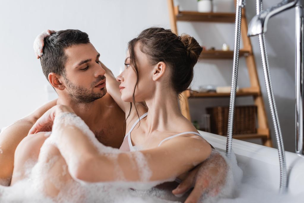 Genç bir kadın banyoda erkek arkadaşına köpükle sarılıp bakıyor.  - Fotoğraf, Görsel