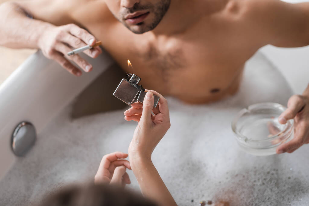 Vista aerea della donna che tiene accendino vicino al fidanzato offuscato con sigaretta e posacenere nella vasca da bagno  - Foto, immagini