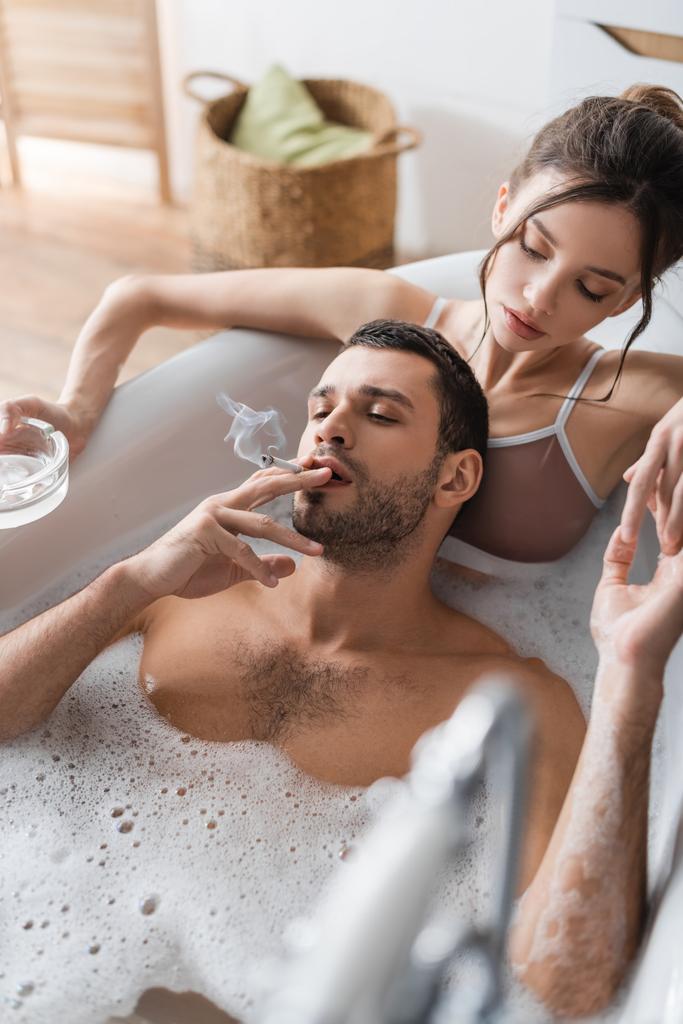 Uomo barbuto fumare sigaretta vicino fidanzata con posacenere nella vasca da bagno  - Foto, immagini