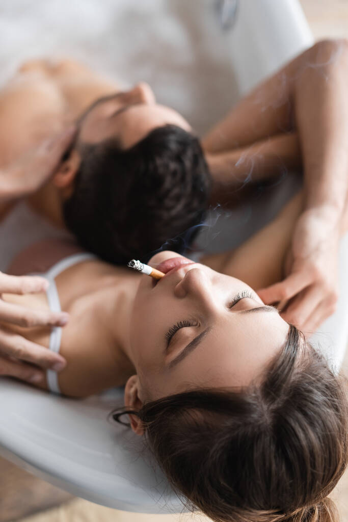 Overhead view of young woman smoking near blurred boyfriend in bath  - 写真・画像