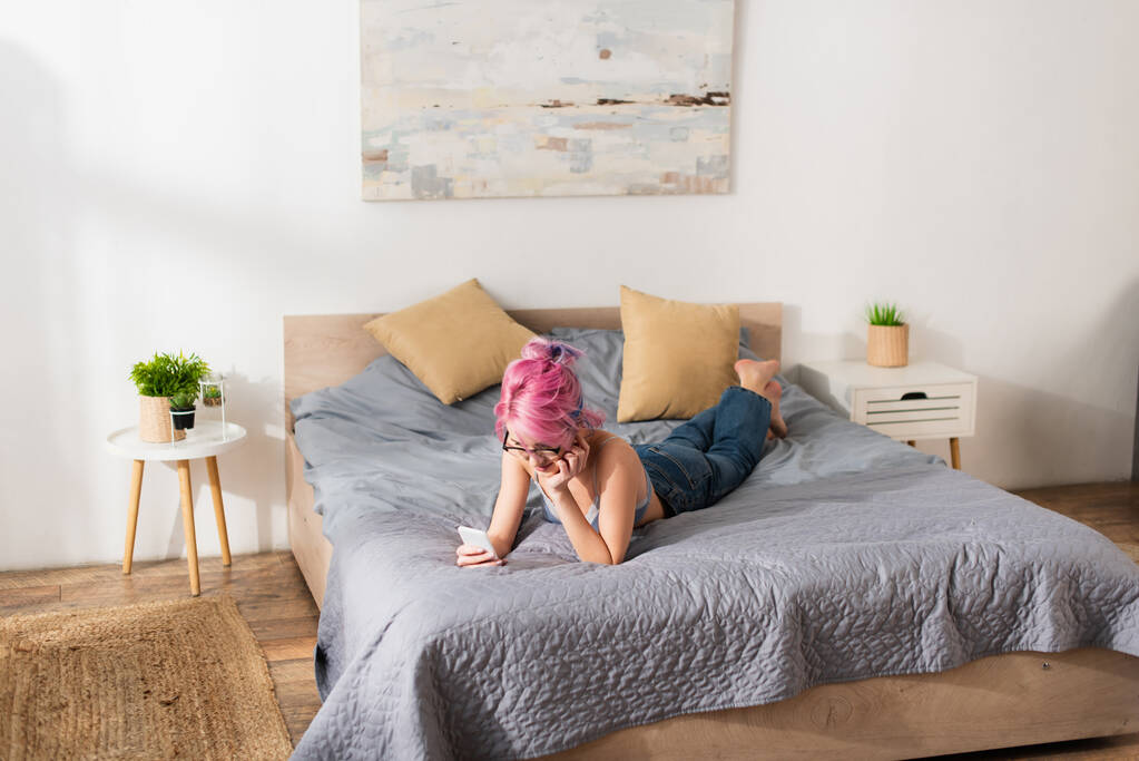 молодая женщина с розовыми волосами в бюстгальтере и джинсах с помощью смартфона во время лежания на кровати  - Фото, изображение