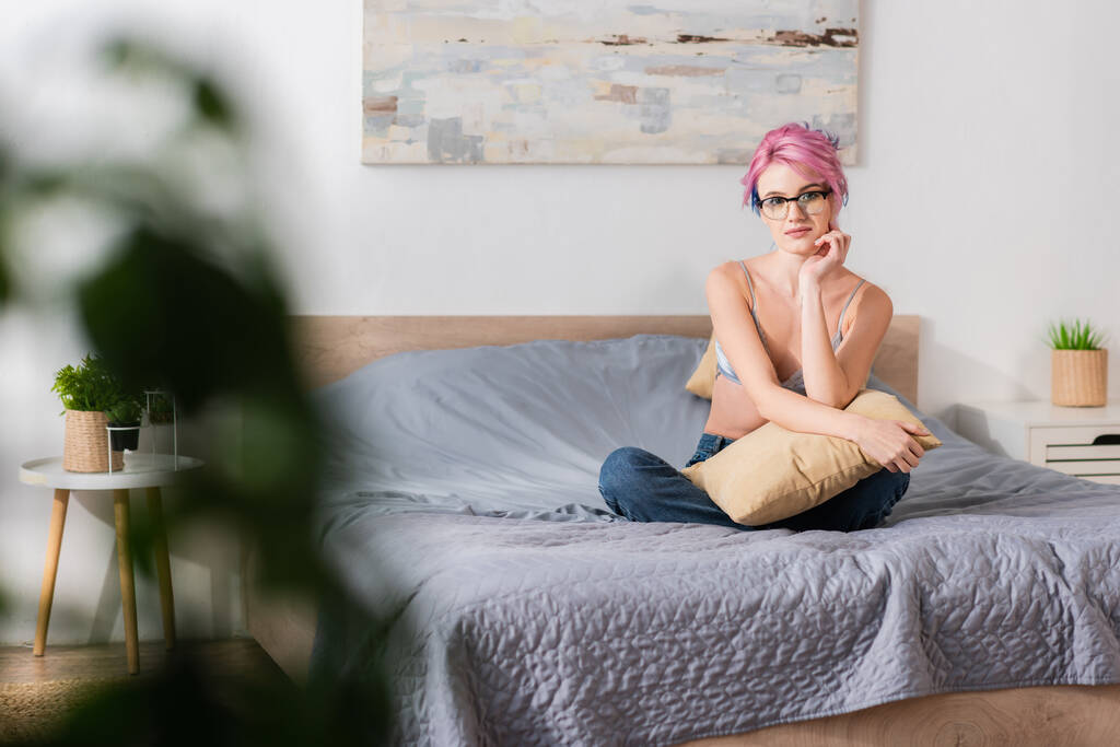 jonge vrouw met geverfd haar zittend in zijden beha en jeans op bed - Foto, afbeelding