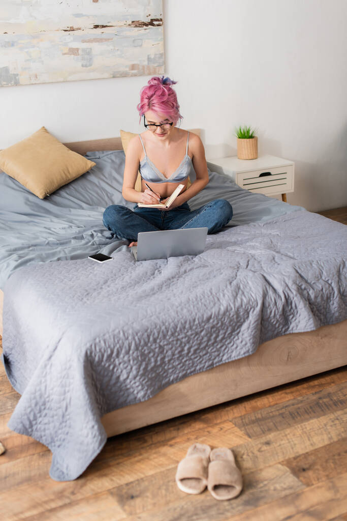 mujer joven con el pelo teñido haciendo notas en el cuaderno mientras estudia en línea en casa - Foto, imagen