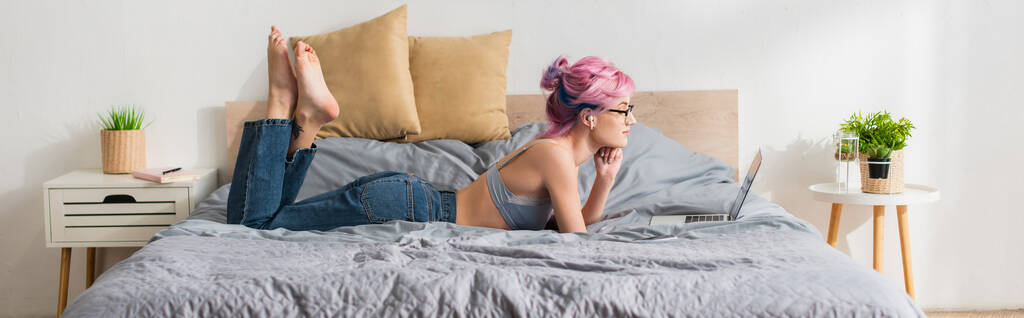 πλαϊνή όψη ξυπόλητης γυναίκας με βαμμένα μαλλιά και ακουστικό ξαπλωμένη στο κρεβάτι και βλέποντας ταινία στο laptop, banner - Φωτογραφία, εικόνα