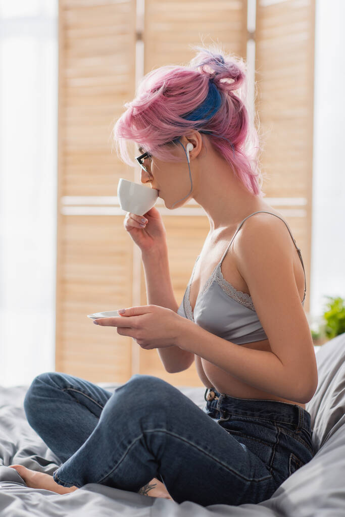 вид сбоку молодой женщины с окрашенными волосами и наушниками, пьющей кофе, сидя на кровати - Фото, изображение