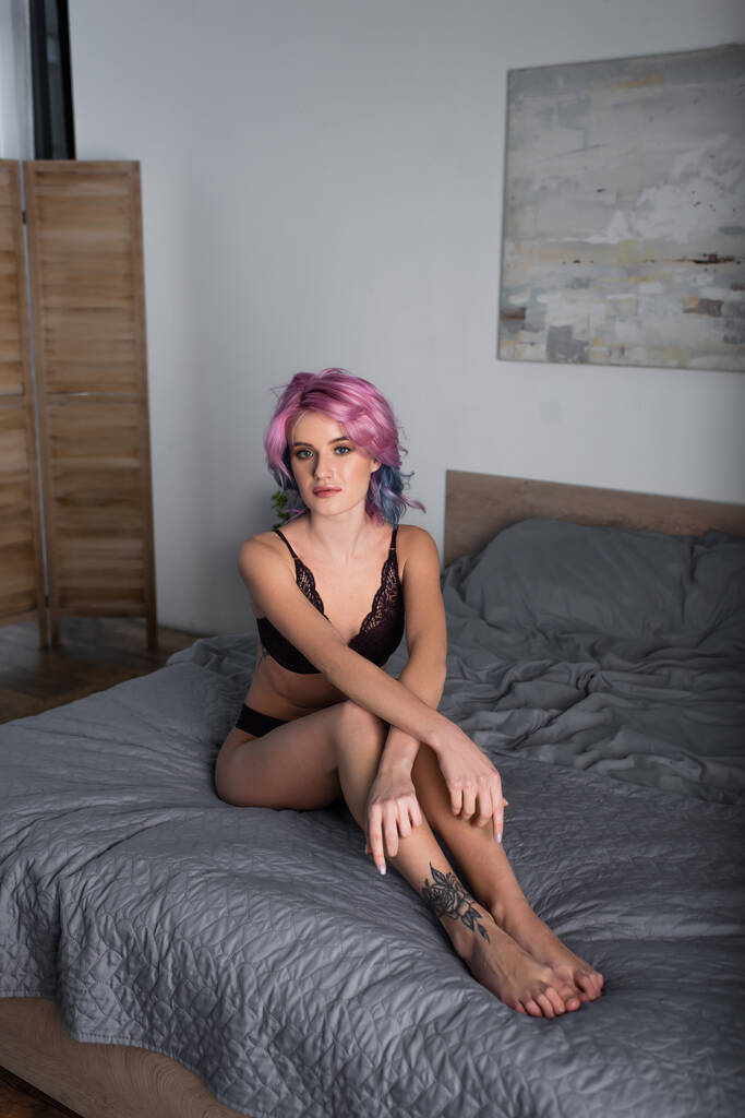 татуированная и сексуальная женщина с окрашенными волосами в темном нижнем белье, сидящая на кровати - Фото, изображение