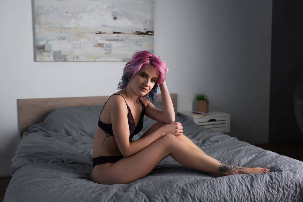 getatoeëerde en sexy vrouw met roze haar in donker kanten ondergoed zittend op bed - Foto, afbeelding