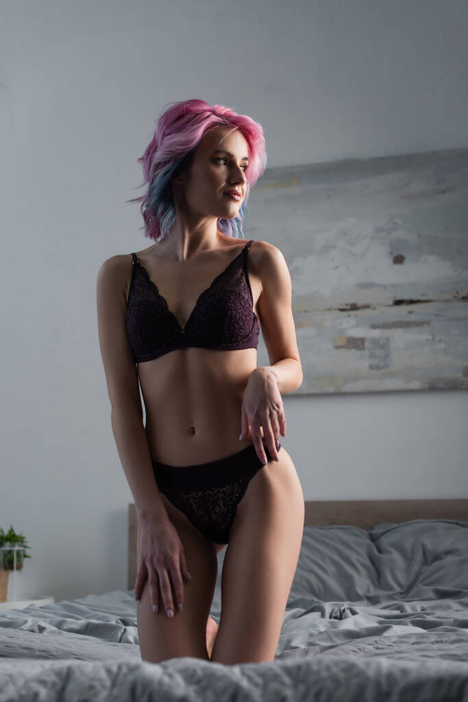 sexy Frau mit gefärbten Haaren in dunklen Dessous posiert zu Hause auf dem Bett - Foto, Bild