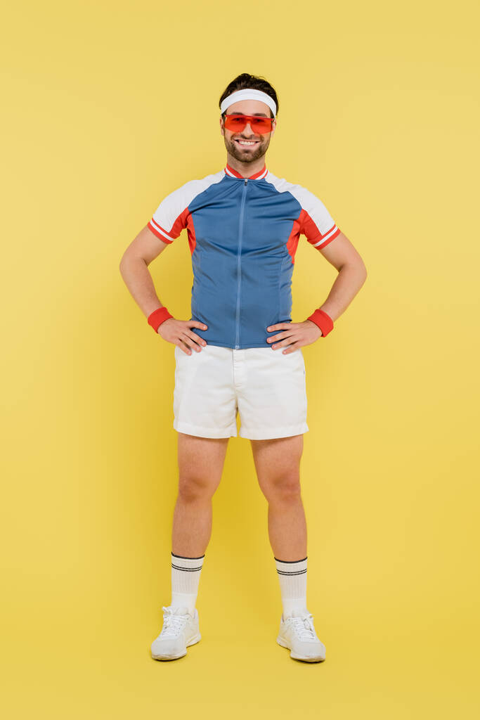Pełna długość uśmiechniętego sportowca w okularach przeciwsłonecznych trzymającego się za ręce na biodrach na żółtym tle  - Zdjęcie, obraz
