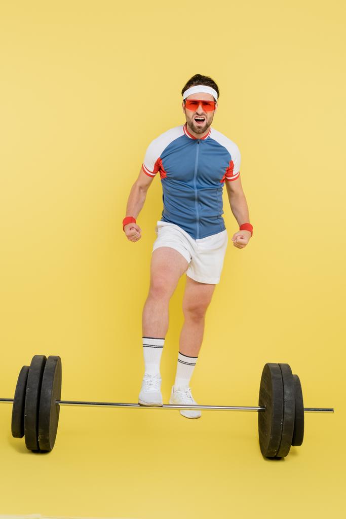 Young sportsman in sunglasses showing muscles near barbell on yellow background  - Photo, Image