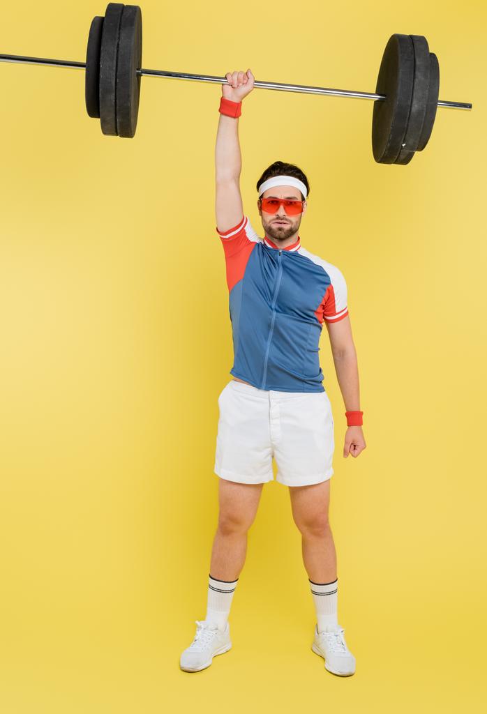 Sportman in zonnebril hijsen lange halter en kijken naar de camera op gele achtergrond  - Foto, afbeelding