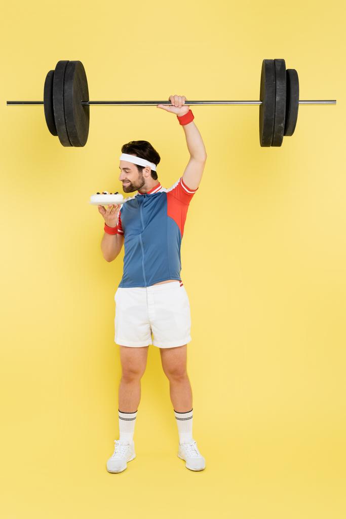 Side view of young sportsman smelling cake and lifting barbell on yellow background  - Photo, Image