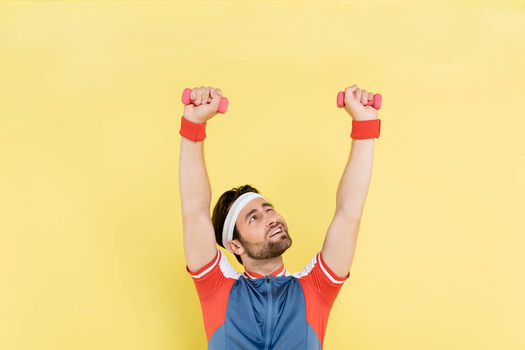 Brunette sportsman lifting dumbbells and looking up isolated on yellow  - Photo, Image