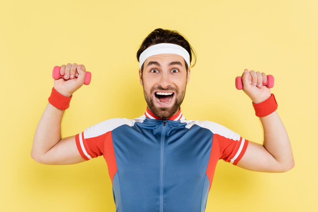 Cheerful sportsman holding dumbbells and looking at camera isolated on yellow  - Photo, Image
