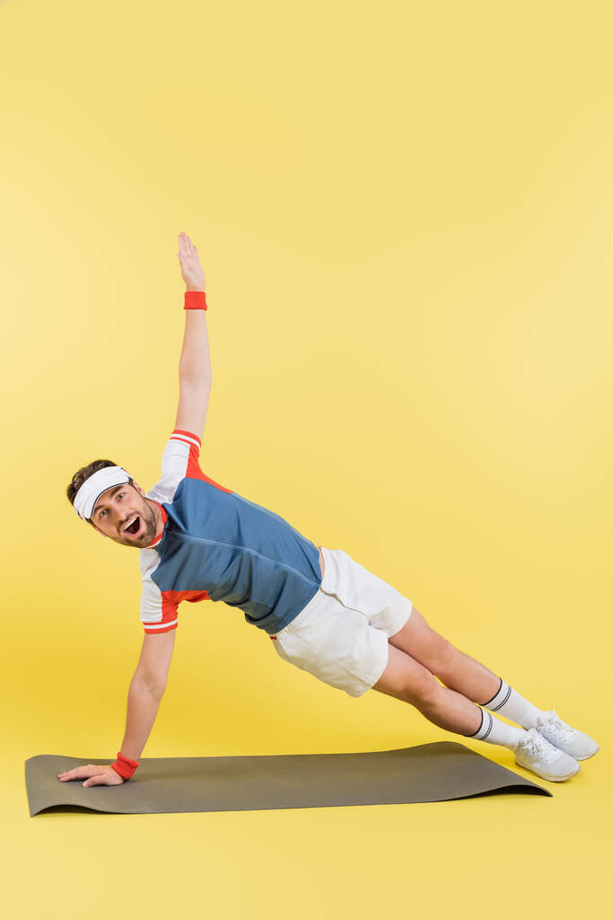 Возбужденный спортсмен в козырьковой тренировке на фитнес-коврике на желтом фоне  - Фото, изображение