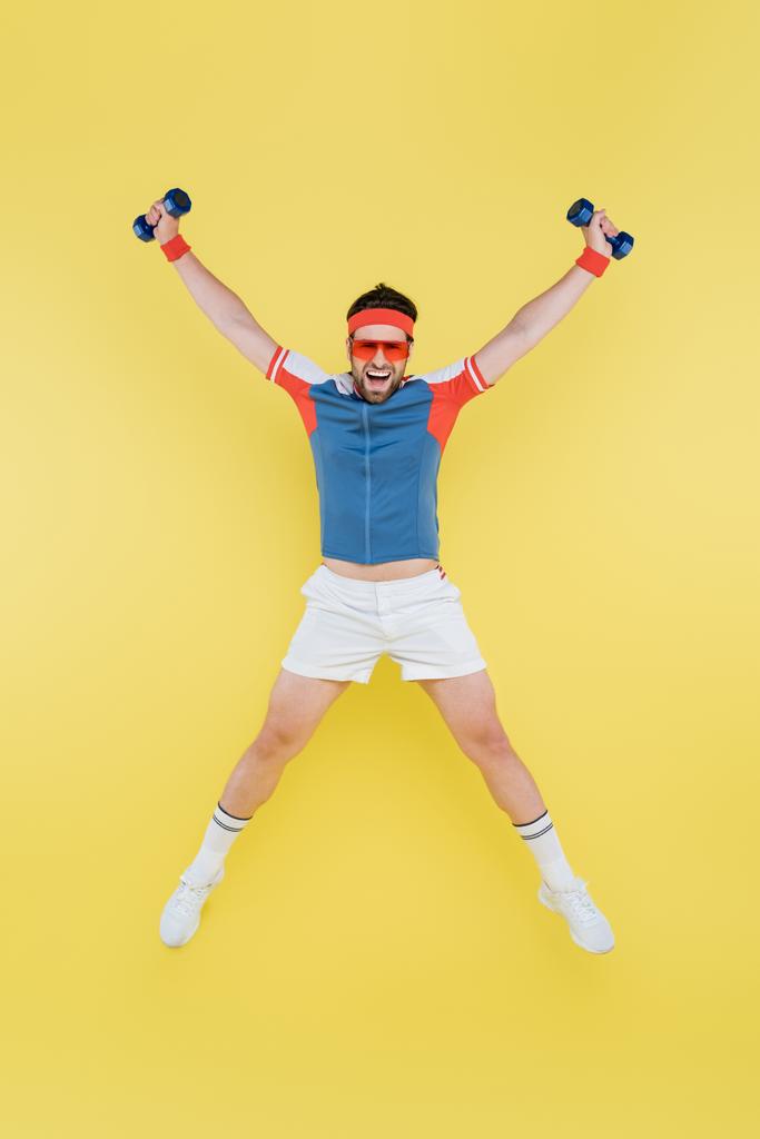 Θετικός αθλητής που κρατάει αλτήρες και πηδάει απομονωμένος στο κίτρινο  - Φωτογραφία, εικόνα