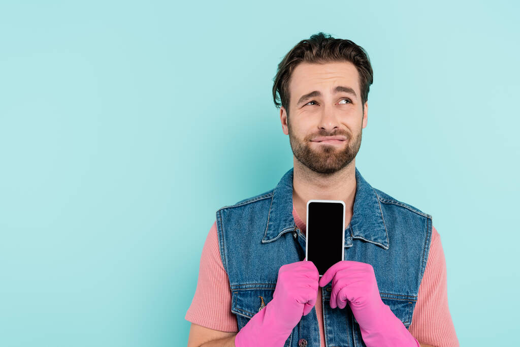 Unzufriedener Mann in Gummihandschuhen hält Handy isoliert auf blauem Grund  - Foto, Bild