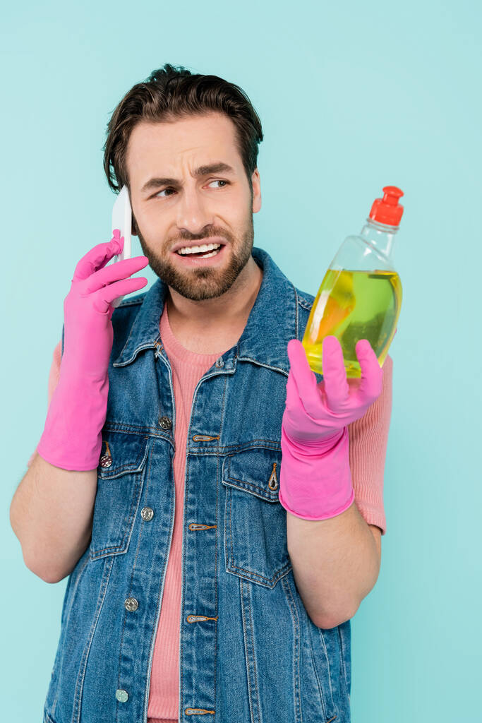 Homme sceptique célibataire en gants en caoutchouc tenant liquide vaisselle et parlant sur smartphone isolé sur bleu  - Photo, image