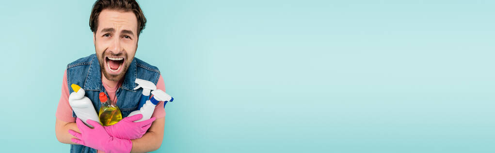 Jovem em luvas de borracha gritando enquanto segurava detergentes isolados em azul, banner  - Foto, Imagem