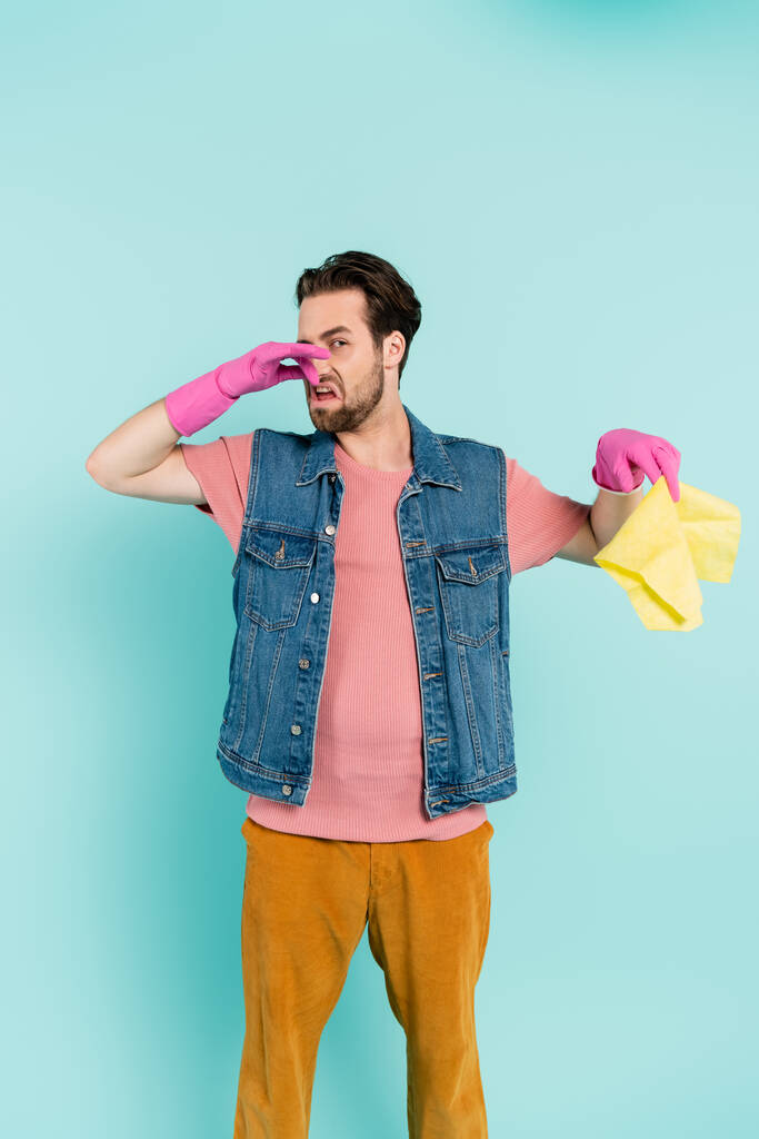 Jeune homme en gants de caoutchouc couvrant le nez tout en tenant tapis isolé sur bleu  - Photo, image