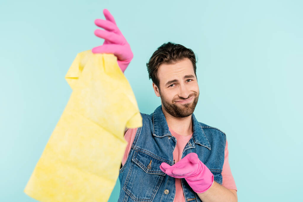 Smiling man in rubber gloves pointing at blurred rug isolated on blue  - 写真・画像