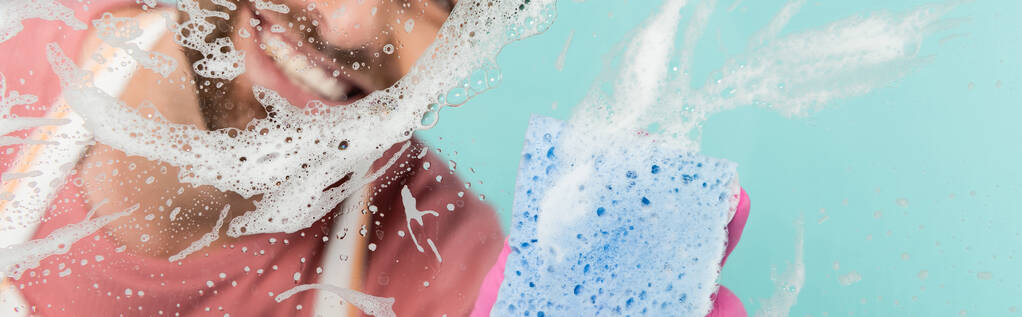 Προβολή του χαμογελαστού ανθρώπου καθαρισμού γυαλί με απορρυπαντικό και σφουγγάρι απομονώνονται σε μπλε, banner  - Φωτογραφία, εικόνα